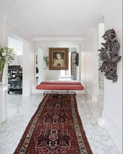 Hall de entrada de um apartamento com piso em marmore, um tapete tipo passadeira persa no chão e um banco vermelho ao fundo.