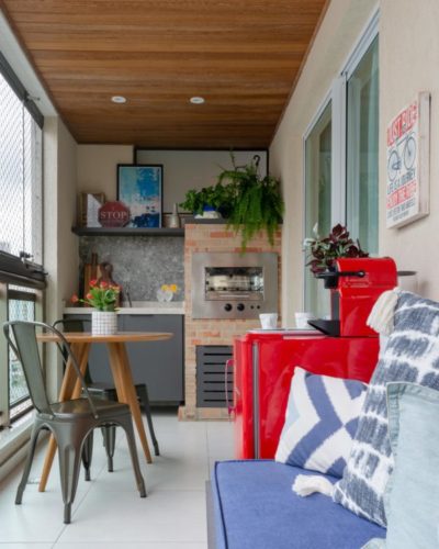 Varanda multiuso, churrasqueira ao fundo, teto revestido em madeira, mesinha redonda e sofá ao lado da mini geladeira vermelha