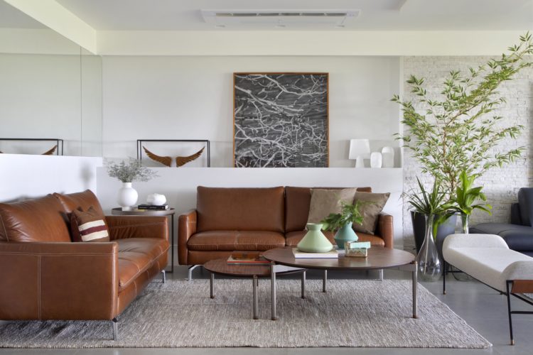 Sala clara, decorada com dois sofás em couro caramelo, duas mesas de centro redondas com alturas diferentes
