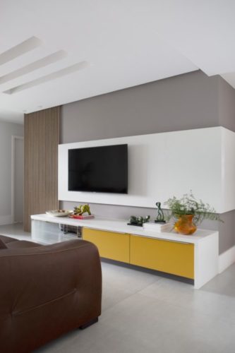 Parede pintada de cinza, um painel em madeira branca no meio para a tv e embaixo um rack branco e amarelo