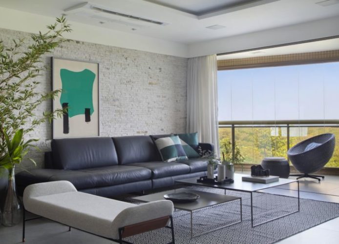 Apartamento na Barra de 288m2 com vista deslumbrante para a Lagoa de Marapendi. Varanda fechada com cortina de vidro, parede de tijolinho branco e sofá em couro azul em frente.