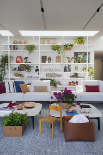 Sala com uma estante branca ao fundo repleto de objetos e na frente um sofá branco com almofadas colorids e tapete azul