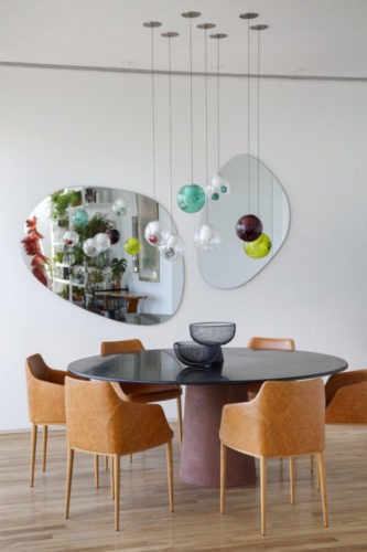 Mesa de jantar redonda, cadeiras em couro, espelhos com formato organico na parede e luminarias pendentes coloridas redondas.