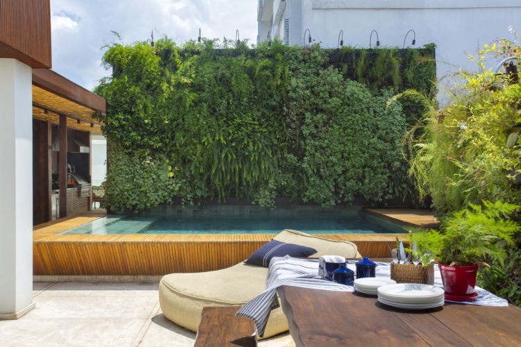 Espaço externo em uma cobertura com piscina, parede verde e uma mesa em madeira.