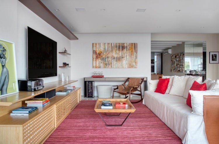 Sala ampla e clara com tapete rosa na fente do sofá branco, rack em madeira baixo e tv em cima