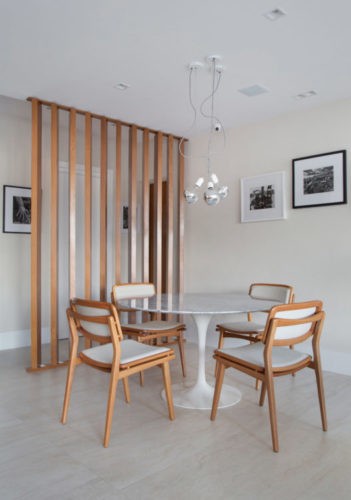Sala de jantar separada da entrada por painel com ripas de madeira, mesa redonda com tampo de marmore e pés branco, com quatro cadeiras em madeira e tecido cru