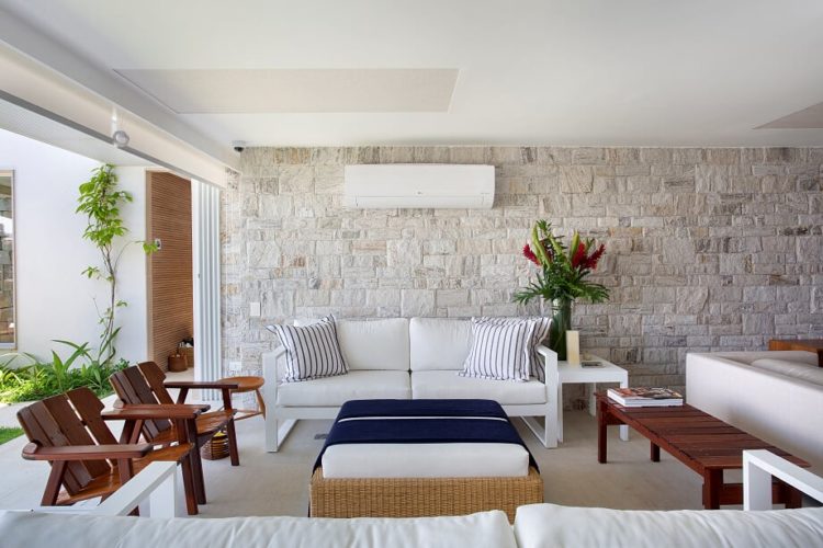 Sala de um casa de praia com parede em pedra, sofá branco e puff de vime em frente
