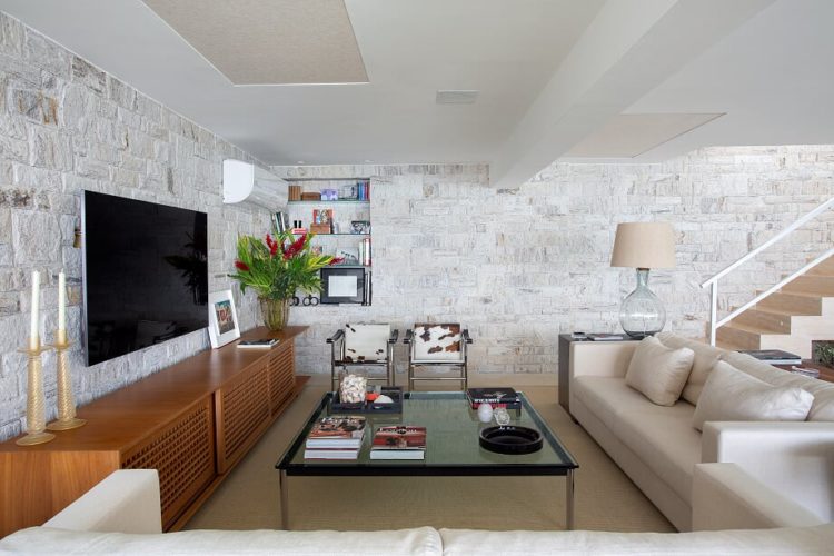 Sala com parede de pedra , rack baixo em madeira, tv na parede e em frente sofá bege