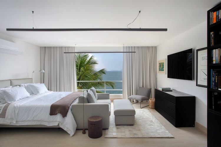 Quarto de casal, cama grande , sofá cinza em frente e de frente para a tv na parede. Sacada aberta com vista para o mar