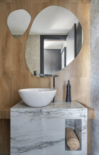 Lavabo com parede revestida em madeira, dois espelhos em formatos orgânicos, bancada em mármore. 
