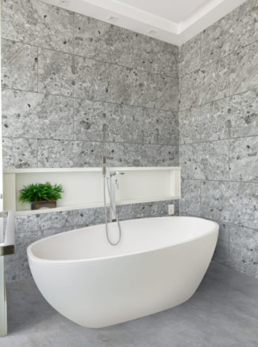 Paredes do banheiro revestida com porcelanato que imita pedra, uma banheira branca no meio.
