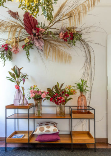 Carrinho de chá com vários arranjos de flores em vasos, e na parede em cima uma arranjo de flores e galhos 