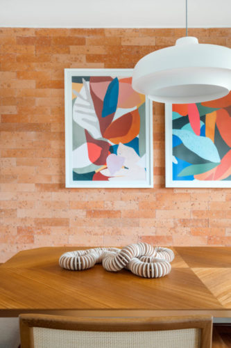 Parede de tijolinho terracota com dois quadros com imagens abstratas coloridas, tampo da mea de jantar em madeira e luminária pendente branca 