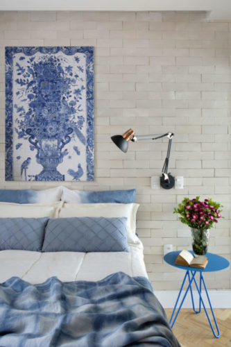 Uma charmosa cobertura no Leme (RJ) com 55m2. Quarto do casal com parede da cama revestida de tijolinho branco, uma painel em azulejos branco e azul com a imagem de um vaso de flor, pendurada em cima da cama Mesinha lateral redonda azul e luminária preta articulável na lateral da cama, instalada na parede 