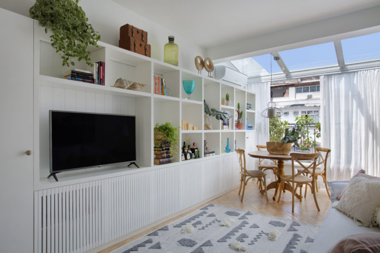 Sala clara, com mesinha redonda em madeira perto da varanda e estante branca para tv e objetos