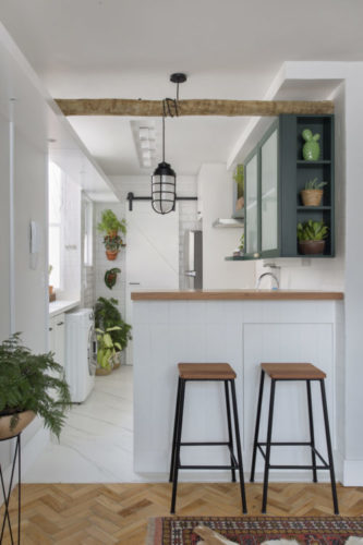 Cozinha branca apenas com os armários superiores na cor verde e cheia de plantas decorando. Um balcão com dois bancos separam da sala 