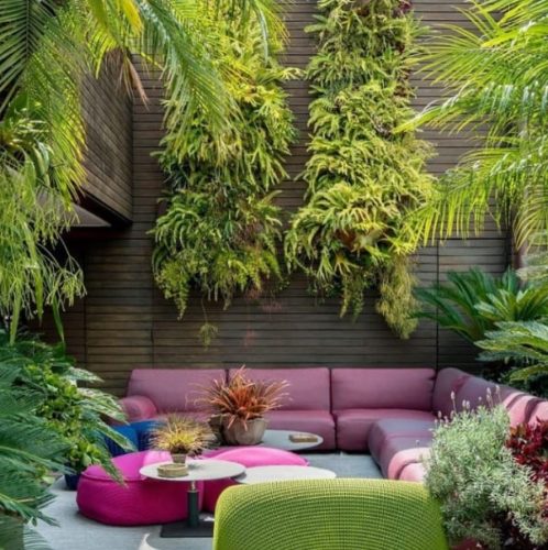 Ambiente externo com uma parede lateral alta e cheia de samambaias, sofá rosa e puffs rosa escuro