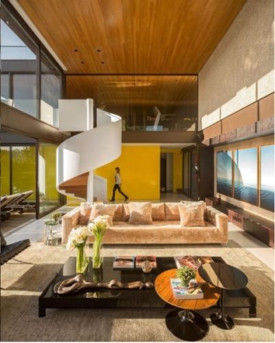O poder das cores nos ambientes. Sala com o pé direito alto, janelas do piso ao teto que é revestido em ripas de madeira, a parede de fundo pintada de amarelo