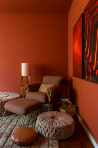 Canto da sala com as paredes pintadas de ocre, uma poltrona ton sur ton e quadros vermelhos na parede