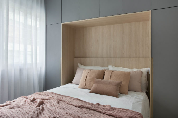 Apartamento compacto - 36m2 muito charmoso, A cama embutida no armário. Parede de fundo do armario revestida em folhas de madeira. Roupa de cama rosa e branca, e as portas dos armários nas laterais e em cima na cor cinza 