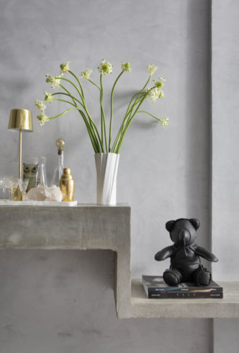 Parede cinza, uma vaso de flor branco com poucas flores, uma bandeja arrumada para bar em cima de uma aparador em concreto e enfeitando um urso preto.