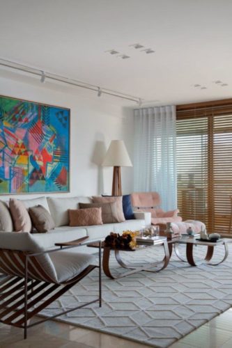 Sala com paredes brancas, persiana horizontal em madeira, cortina de voal ao lado, sofá bege e em cima do sofá, tela colorido de Burle Marx