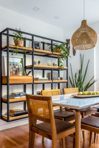 Sala de jantar com mesa e cadeiras em madeira, luminária pendente em palha e na parede, uma estante suspensa em ferro e madeira com objetos para enfeitar, como livros e plantas