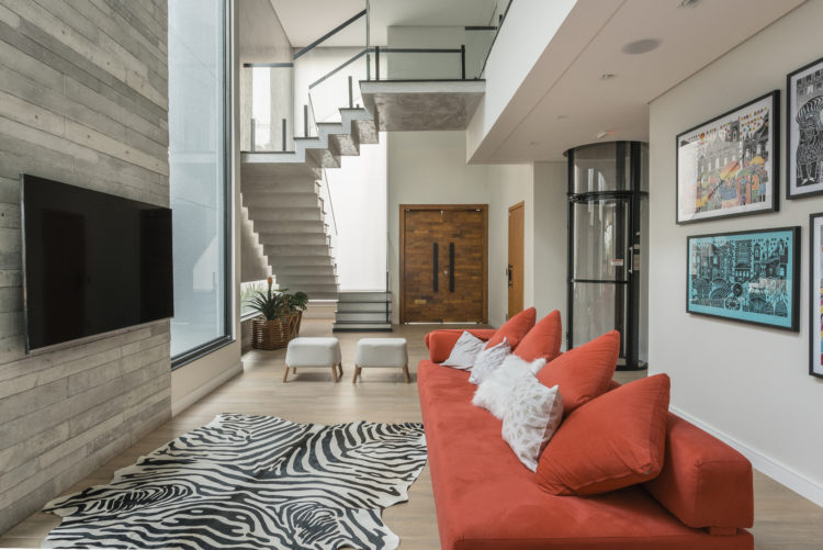 Decoração em diversos estilos nessa living com sofá vermelho, tapete de zebra e painel de porcelanato imitando madeira. Ao fundo escada em mármore, porta de entrada em madeira e ao lado elevador circular aparente.