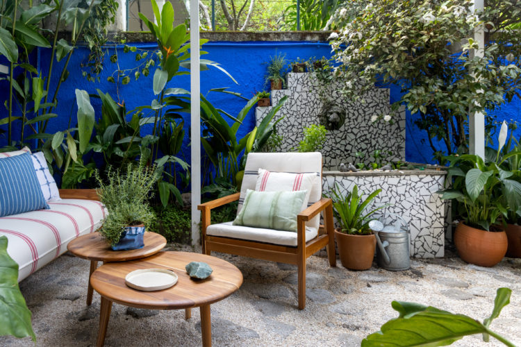 Espaço externo com o muro lateral pintado de azul, fonte revestida com cacos de cerâmica, moveis em madeira ambientado