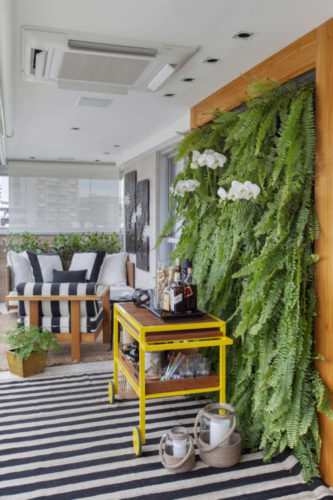 Varanda com parede viva de plantas, com samambaias, carrinho amarelo em frete, usado com bar, tapete listrado e ao fundo, sofá listrado