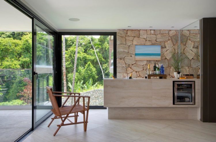 Espaço do bar na cobertura, ao fundo parede de pedra. Na frete bancada em mármore com geladeira de vinho embutida, janelões integram a natureza. 