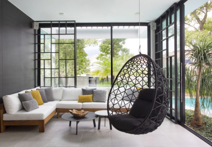 Sala de estar, com vidros , integram a paisagem da piscina, Sofá em L branco e uma cadeira de balanço suspensa preta.