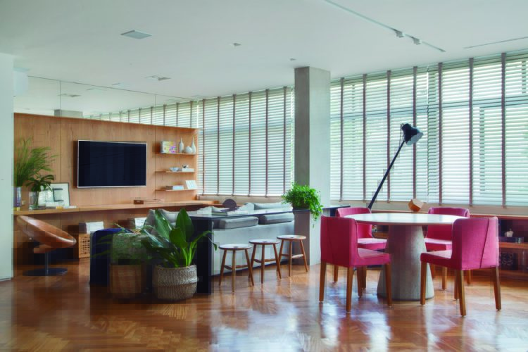 Ampla sala de estar, janelões com persianas na horizontal, parede revestida de madeira ao fundo, para abrigar a TV, no primeiro plano, mesa redonda com quatro cadeiras rosas.
