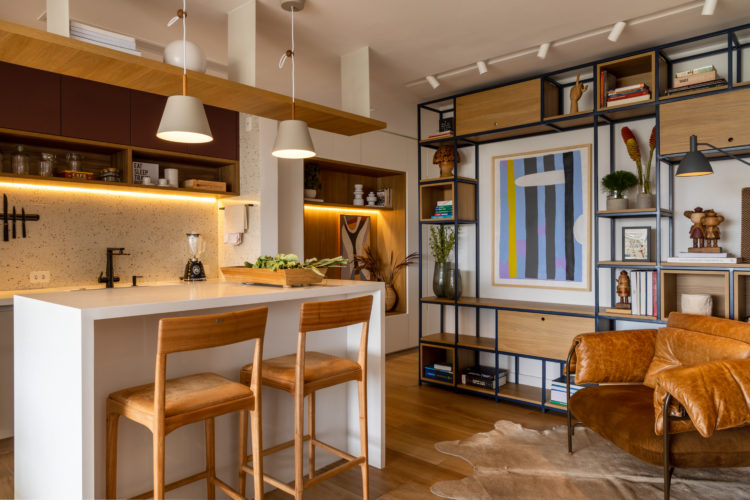 Varanda integrada no apartamento de 90m² na Zona Sul carioca. Cozinha também integrada, estante em ferro e poltrona em couro