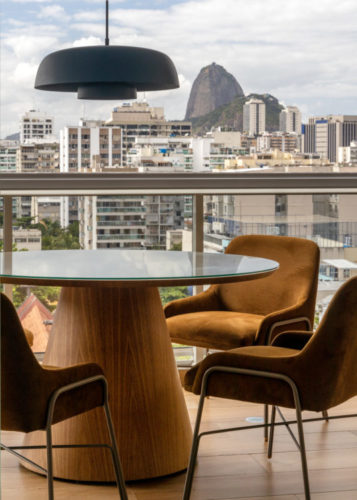 Varanda integrada no apartamento de 90m² na Zona Sul carioca, com vista para o pão de açúcar ao fundo. Mesa redonda com pé central, cadeiras em couro marrom e pendente na cor azul no centro