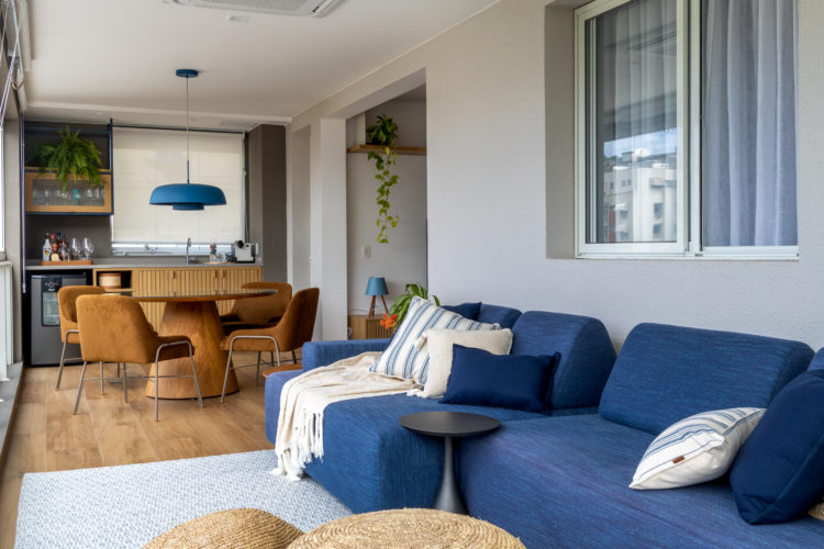 Varanda integrada no apartamento de 90m² na Zona Sul carioca. Sofá azul, ao fundo mesa de jantar redonda com cadeiras em couro, bancada com cuba e mini geladeira