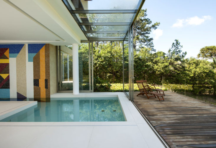 Varanda com deck em madeira, atrás, piscina coberta mas integrada por grandes portas de correr em vidro, e na parede fundo da piscina um painel de azulejos de Noel Marinho.