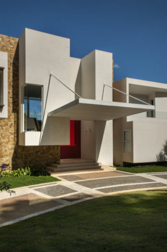 Último projeto residencial do arquiteto e urbanista Noel Marinho. Entrada da casa, volume branco em forma de retângulo na vertical, porta de entrada vermelha e um marquise quadrada em balanço.