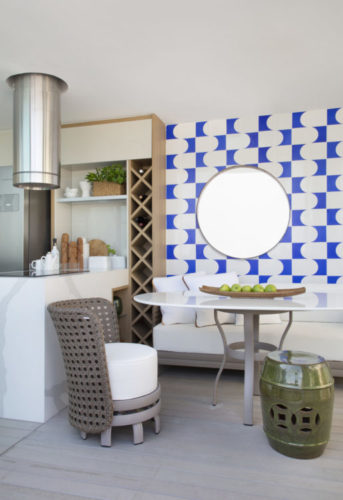 Varanda fechada com espaço gourmet, coifa, churrasqueira e cooktop na bancada. Ao lado mesa com sofá e na parede atrás um painel em azulejos branco e azuis