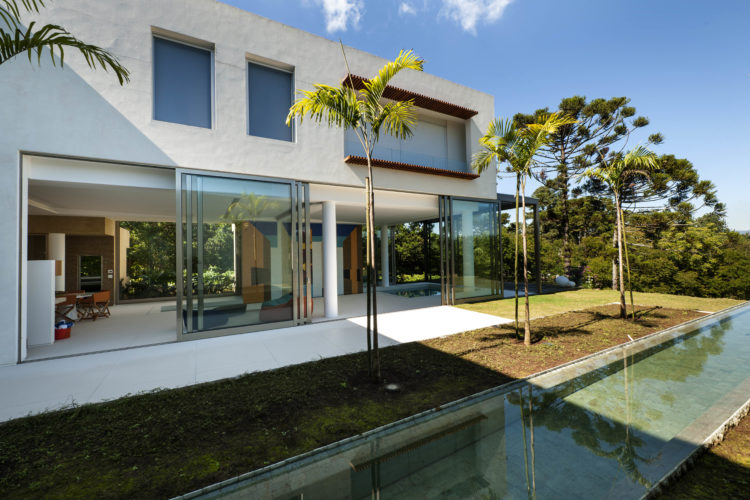 Casa com fachada branca, grandes portas de correr em frete a um jardim com raia de piscina e palmeiras