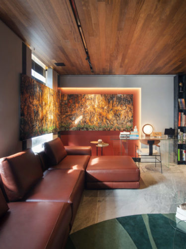 Sofá em couro marrom com chaise, na parede lateral, um nicho iluminado e pintado de laranja abriga um quadro.
