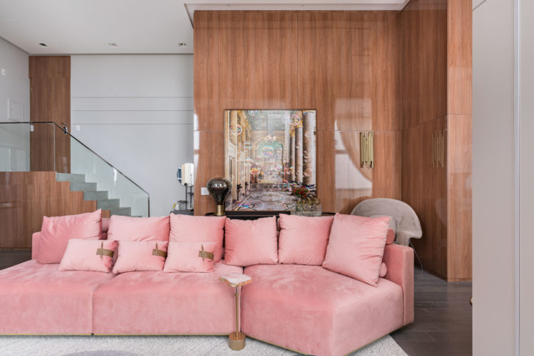 Decoração chic e contemporânea, sofá curvo na cor rosa no centro da sala. Ao fundo parede revestida em painel de madeira brilhoso 