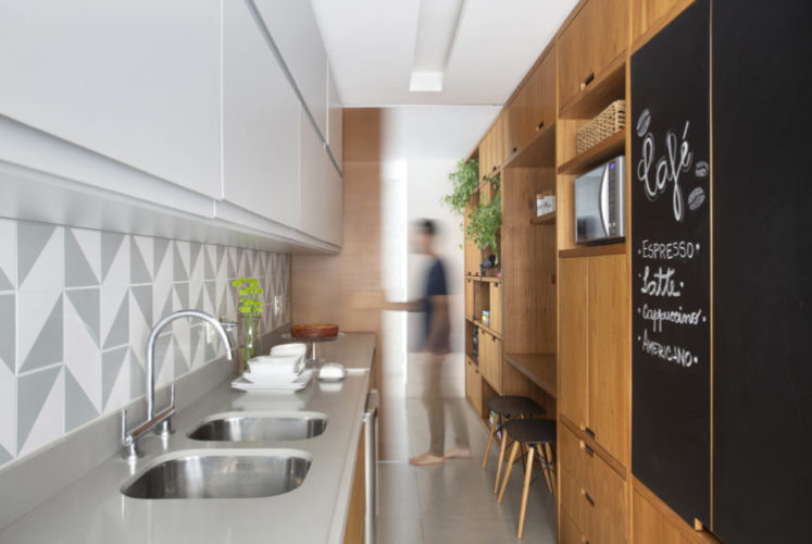 Cozinha com planta em corredor, ao fundo, porta de correr em madeiro freijó para integrar ambientes. Armários da cozinha na mesma madeira. E detalhe de uma porta em lousa 