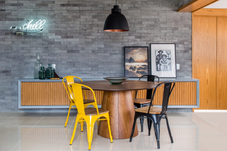Décor com pegada industrial na área social, parede revestida com tijolinho cinza, mesa redonda em madeira, com cadeiras amarelas e pretas, portas de correr em madeira