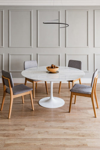 Ambiente da sala de jantar com molduras em madeira aplicadas na parede (boiserie) pintadas na mesma cor bege das paredes. Mesa em mármore redonda e quatro cadeiras.