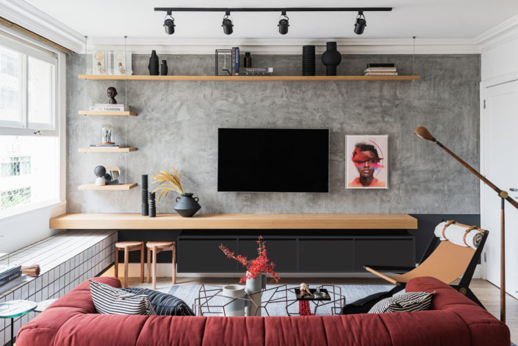 Décor da área social em um apartamento alugado. Parede pintada de cinza, tv na parede, embaixo uma pratelira em madeira com móvel baixo preto.