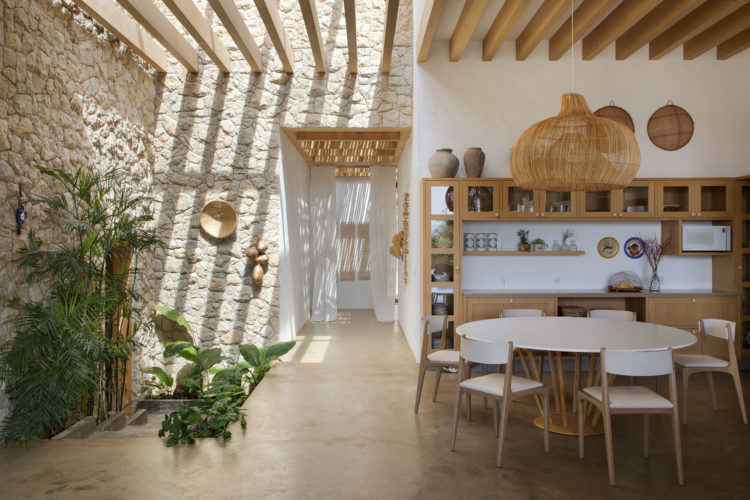 Sala de uma casa de veraneio, com paredes de pedra, iluminação zenital com ripas em madeira, cozinha integrada