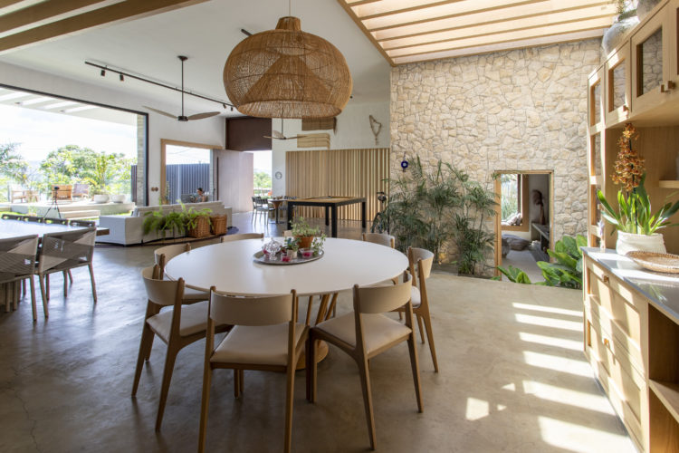 Sala de uma casa de veraneio com todos os espaços integrados, piso em cimento, parede de pedra, mesa redonda e ao fundo , área externa 