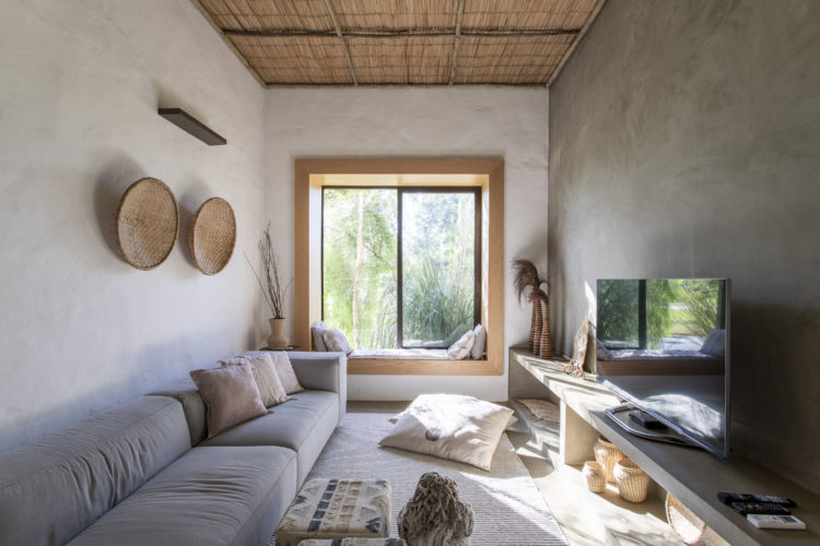 Sala de tv de uma casa de veraneio, piso e paredes em cimento, ao fundo, janela com banquinho na sua extenção