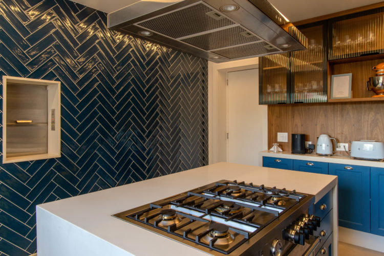 Cozinha com uma parede revestida de azulejo modelo metrô na cor azul e instalado na paginação chevron, no meio um monta carga. Ilha com fogão e coifa 